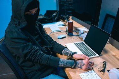 busted hacker in mask with handcuffs in front of workplace clipart