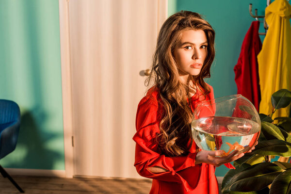 stylish woman in red dress holding aquarium with gold fish and looking at camera at home