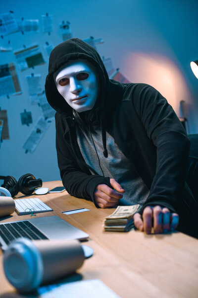 hacker in mask with stack of money on desk