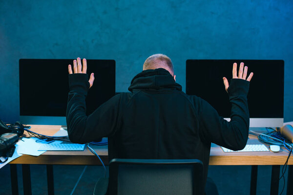 Задний вид арестованного хакера с поднятыми руками перед рабочим местом
