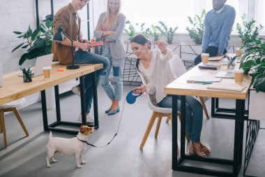 businesswoman playing with jack russel terrier on leash and colleagues standing behind in modern office