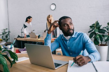 Tablo ve arkasında modern ofisinde çalışan iş arkadaşlarınızla oturup düşünceli Afrikalı-Amerikalı iş adamı 