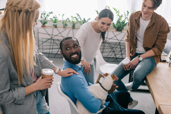 smiling african american businessman sitting with dog surrounded by colleagues with paper cups of coffee 