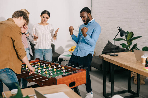 african american celebrating victory in table football and colleagues standing near 