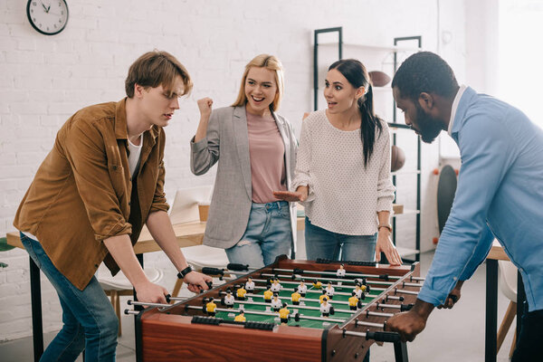 side view of multicultural businessmen playing table football in front of female colleagues 