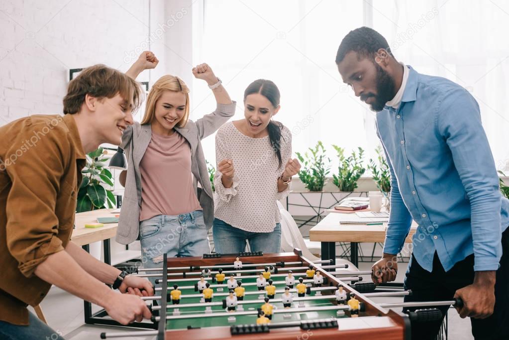 side view of happy multicultural businessmen playing table football in front of celebrating and gesturing female colleagues 