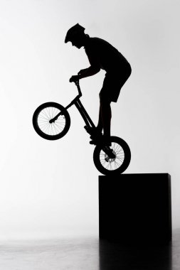 silhouette of trial biker performing stunt while balancing on cube on white clipart