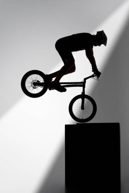 silhouette of trial cyclist performing stunt while balancing on cube on grey clipart