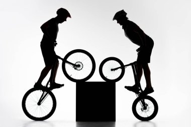 silhouettes of trial bikers performing stunt with cube synchronously on white clipart