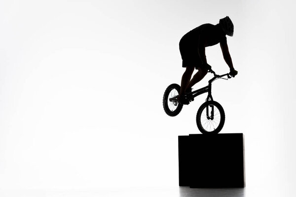 silhouette of trial cyclist performing stunt while balancing on cube on white