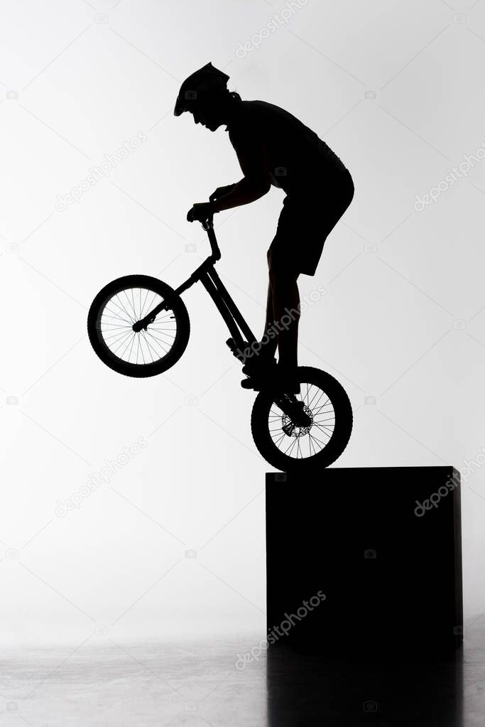 silhouette of trial biker performing stunt while balancing on cube on white