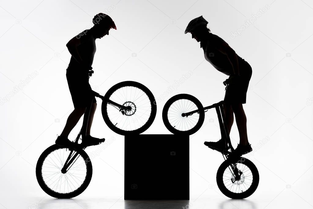 silhouettes of trial bikers performing stunt with cube synchronously on white