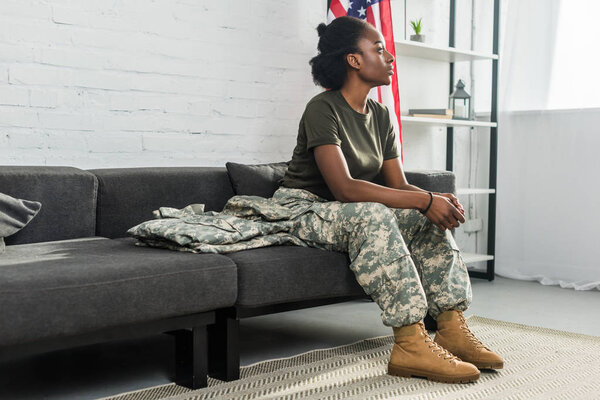 Young female soldier in camouflage clothes waiting on sofa in room