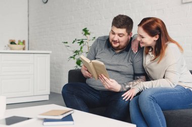 boyfriend and smiling girlfriend reading book together on sofa in living room clipart