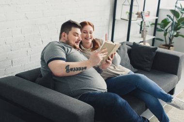 Smiling overweight boyfriend and girlfriend reading book on sofa at home clipart