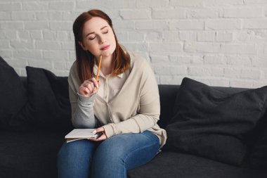 pensive beautiful girl with closed eyes touching chin with pencil at home clipart