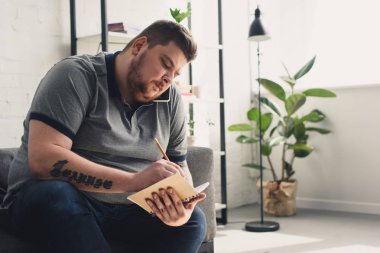 Handsome overweight man writing something to notebook on sofa at home clipart