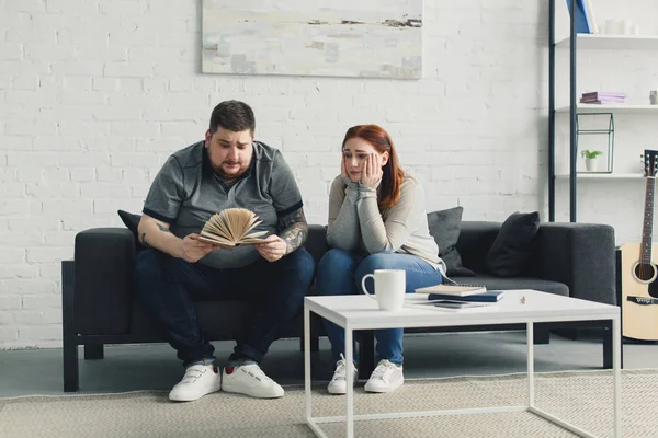 boyfriend searching something in book, sad girlfriend sitting near on sofa