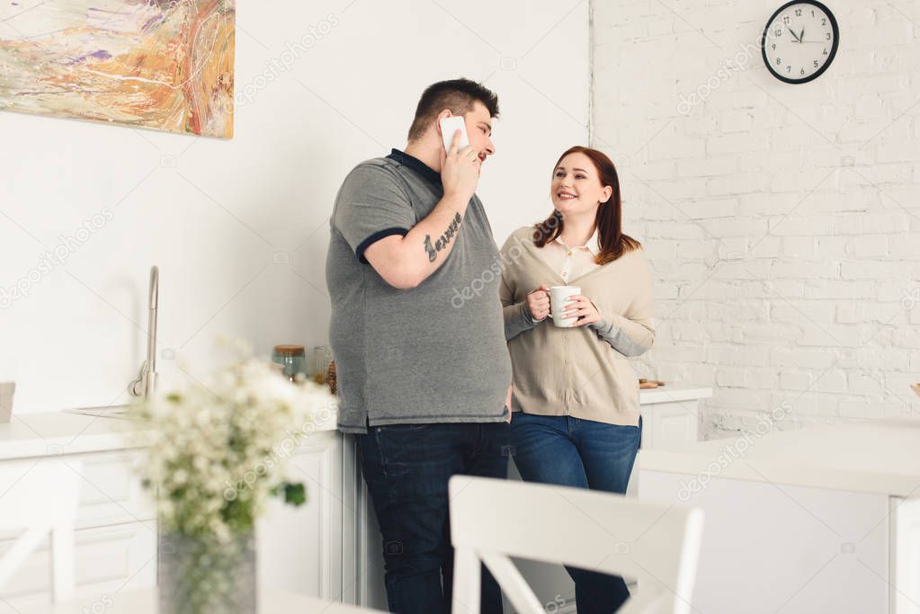 boyfriend talking by smartphone and happy girlfriend holding cup of coffee in kitchen