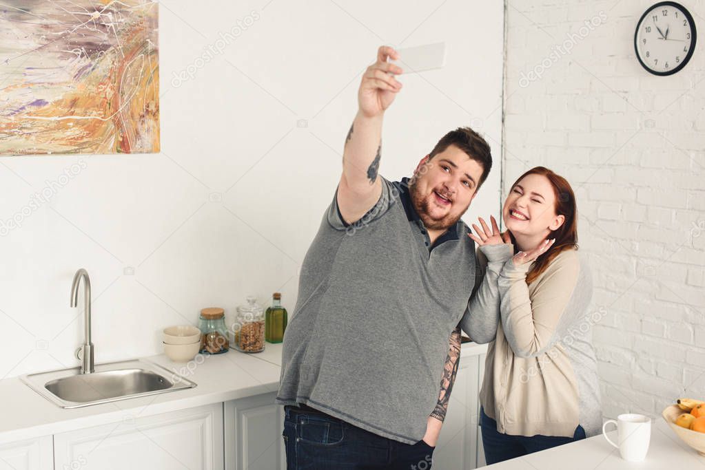 smiling boyfriend and girlfriend taking selfie with smartphone in kitchen