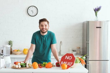 smiling handsome vegan man standing near kitchen counter with vegetables and no meat sign clipart