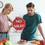 Vegan couple with no meat sign at table with raw meat and vegetables in kitchen at home