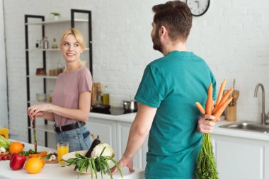 vegan boyfriend hiding bouquet of carrots from girlfriend at kitchen clipart