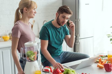 vegan yaşam tarzı konsept dizüstü bilgisayar ve kız arkadaşı yakınındaki tarafından evde, mutfak masasında salatalık konuşurken gibi davranarak adam