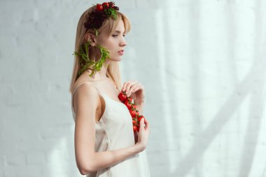 side view of young woman with cherry tomatoes in hands, wreath made of fresh vegetables and earring made of fresh arugula, vegan lifestyle concept clipart