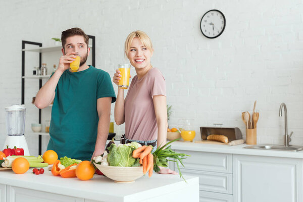 smiling couple of vegans drinking fresh juice at kitchen and looking away
