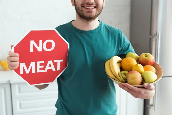 partial view of smiling man holding no meat sign and bowl with fresh fruits, vegan lifestyle concept