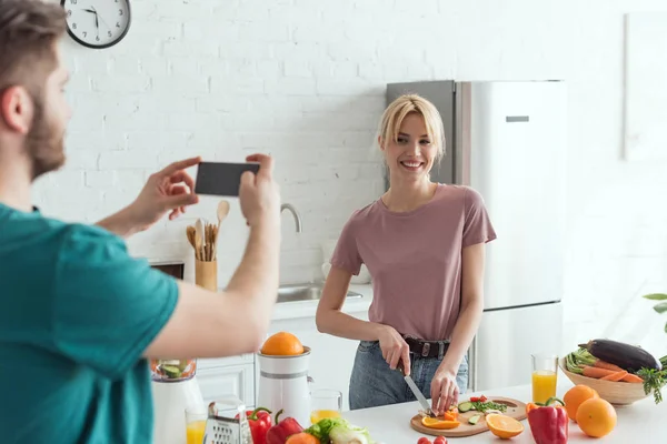partial view of man taking picture of smiling girlfriend cooking in kitchen at home, vegan lifestyle concept