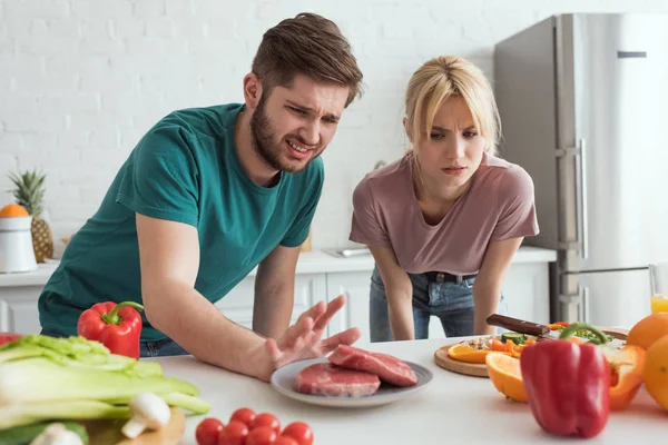 disgusted vegan couple looking at raw meat on plate in kitchen at home
