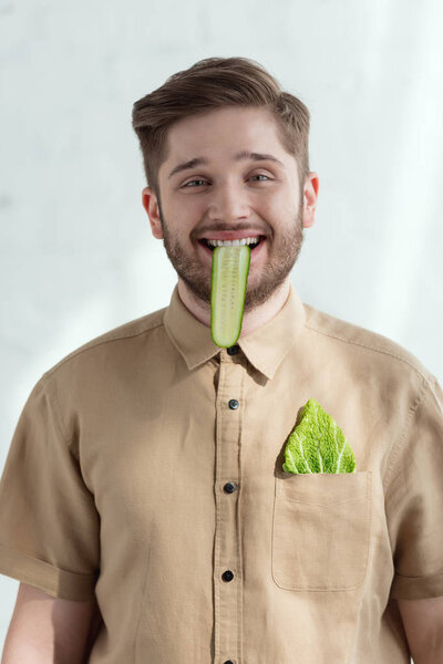 portrait of smiling man with cucumber slice in mouth and savoy cabbage leaf in pocket, vegan lifestyle concept 