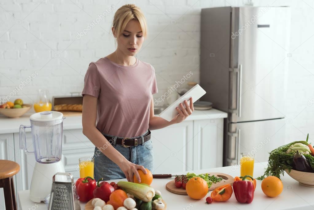 attractive vegan girl cooking and holding tablet with recipe in kitchen