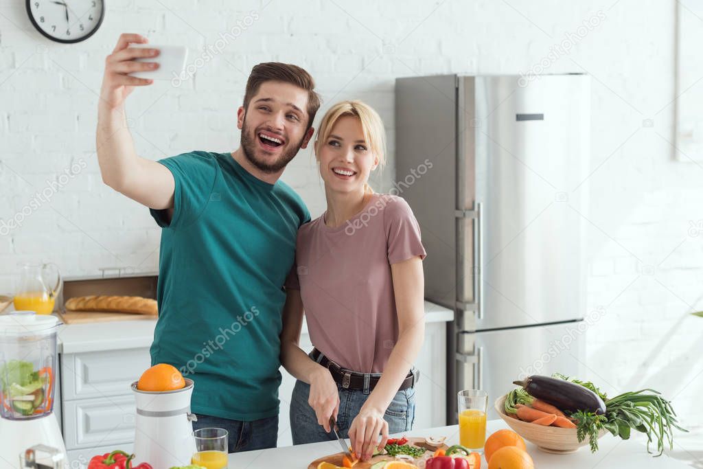 smiling vegan couple taking selfie while cooking together in kitchen at home