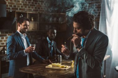 handsome young businessman smoking cigar while smiling friends drinking alcohol beverages behind  clipart