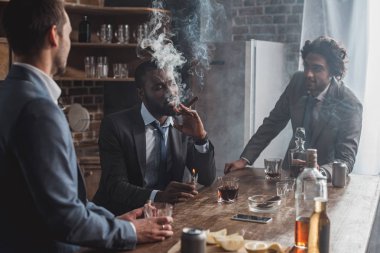 young multiethnic businessmen talking while smoking cigars and drinking whiskey clipart