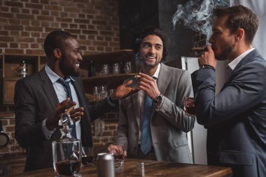 smiling multiethnic male friends in suits talking, drinking whiskey and smoking cigars clipart