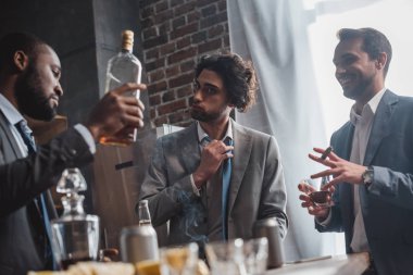 low angle view of young multiethnic businessmen smoking cigars and drinking whisky clipart
