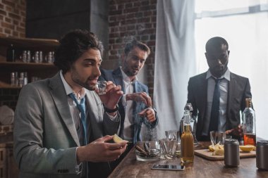 multiethnic male friends in formal wear drinking tequila and smoking cigars clipart