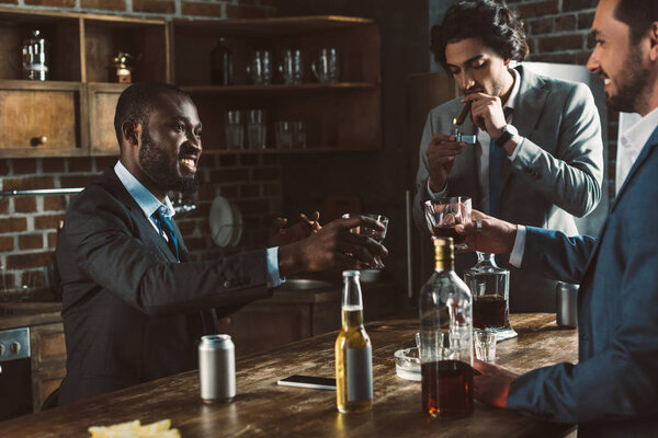 обрезанный снимок улыбающихся многонациональных друзей-мужчин в костюмах, пьющих алкогольные напитки и курящих сигары вместе
