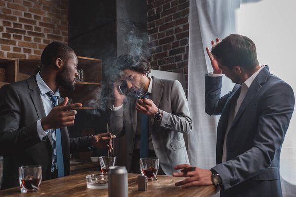 emotional young businessman smoking cigar and talking by smartphone while friends drinking whiskey