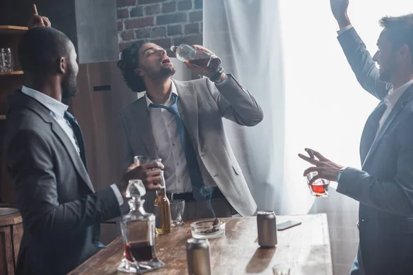 happy multiethnic friends in suits drinking alcohol and smoking cigars together