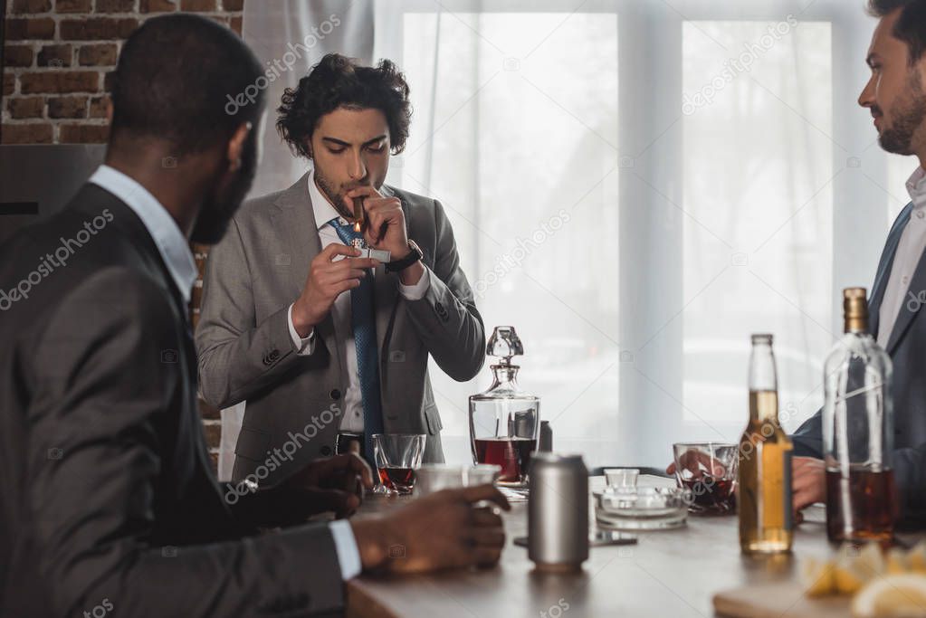 cropped shot of multiethnic men drinking alcohol and looking at friend smoking cigar