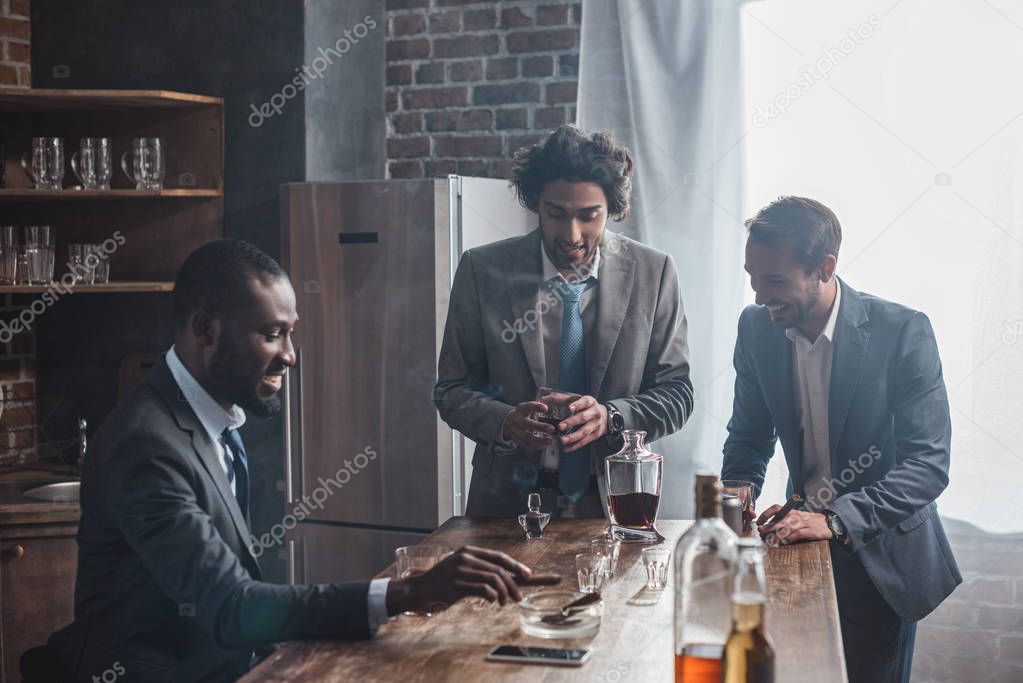 smiling young multiethnic male friends in formal wear drinking whiskey and smoking cigars together