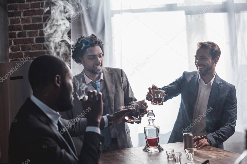 three smiling multiethnic businessmen smoking cigars and clinking glasses of whiskey