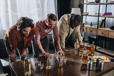 young multiethnic men looking at glasses with alcohol beverages while partying indoors clipart