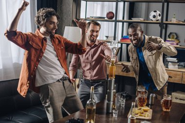 cheerful multiethnic men friends having fun and looking at tower from bottles and glasses while partying indoors clipart