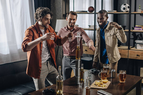 веселые друзья мужского пола смотрят на башню из бутылок и стаканов во время вечеринок в помещении
